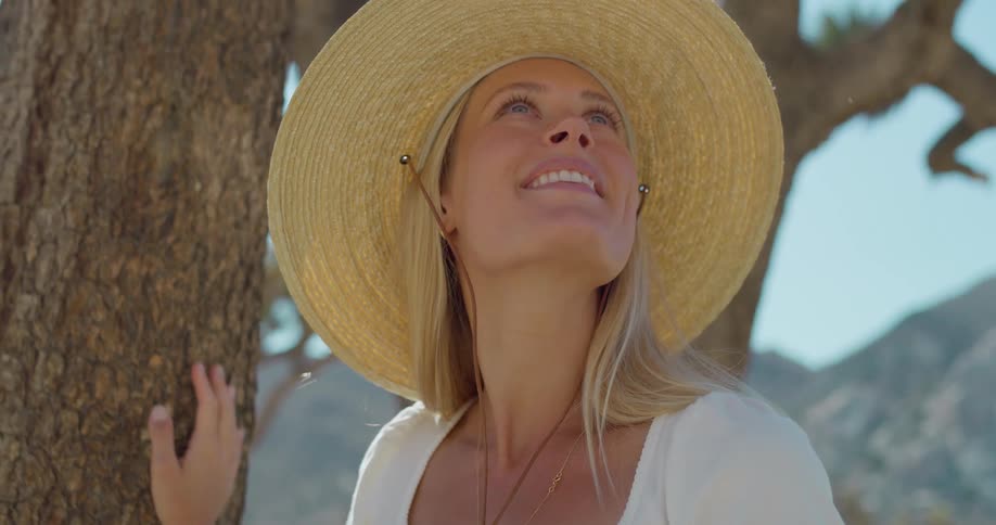 A blonde woman wearing a widebrimmed straw hat and a white blouse stands next to a gnarled tree she looks up and smiles basking in the sun