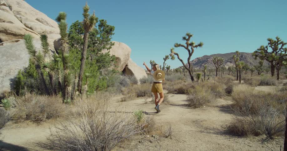 A woman wearing a straw hat and a skirt walks through a desert landscape filled with joshua trees and large rock formations