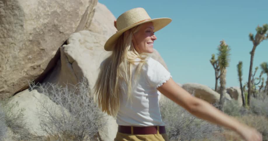 A woman with long blonde hair and a straw hat walks away from the camera through a desert landscape with large boulders turns points off screen and smiles