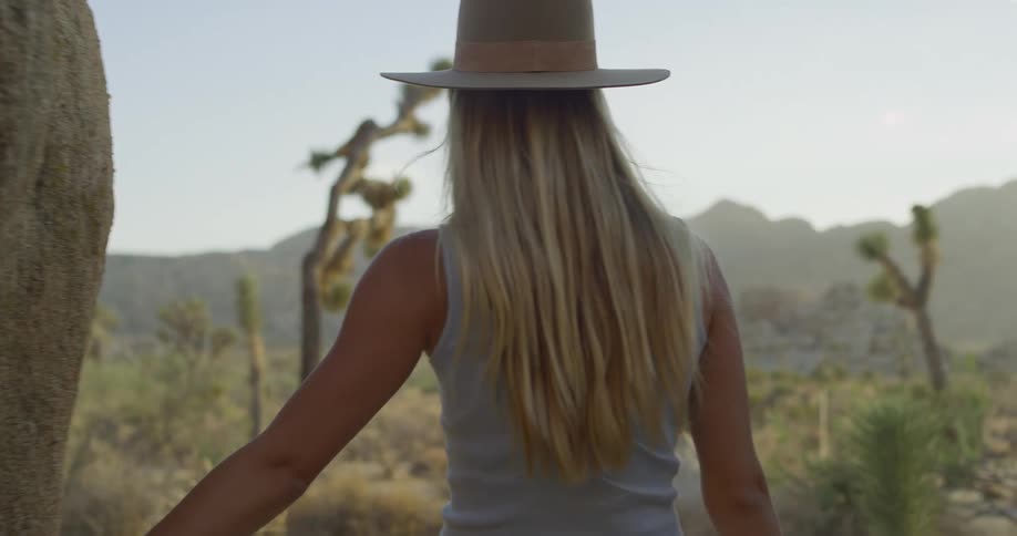 A blonde woman with her back to the camera wearing a hat is hiking in a rocky desertlike landscape the area is filled with large boulders and joshua trees under a bright hazy sky