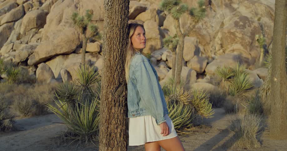A blonde woman wearing a white dress and a denim jacket is standing next to a tree in a desert landscape she leans against the tree and looks at the camera before turning her gaze to the side