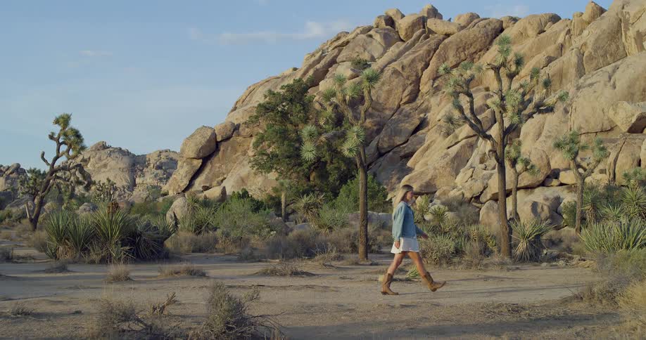 A young woman walking on a barren land wearing a jean jacket and boots at midday with a large rock in the background