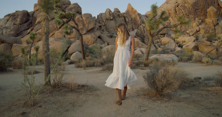 A young woman wearing a white gown and brown boots walks on rocky terrain at midday