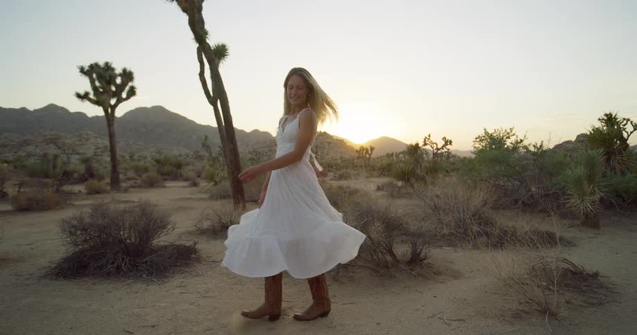 The video shows a wide shot of a young woman standing in a desert landscape during sunset she is wearing a flowing white dress and cowboy boots and she smiles as she twirls around