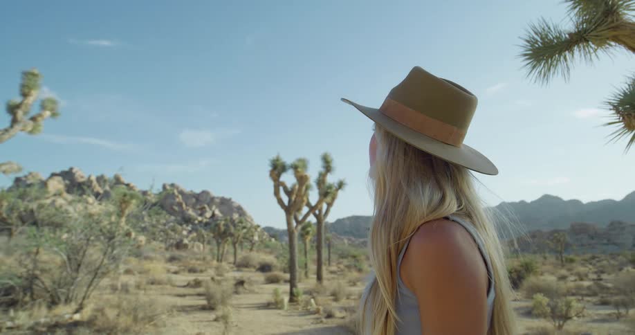 A woman with long blonde hair and a beige hat stands in a desert with joshua trees and large rocks looking up at the clear blue sky before looking around with a neutral expression