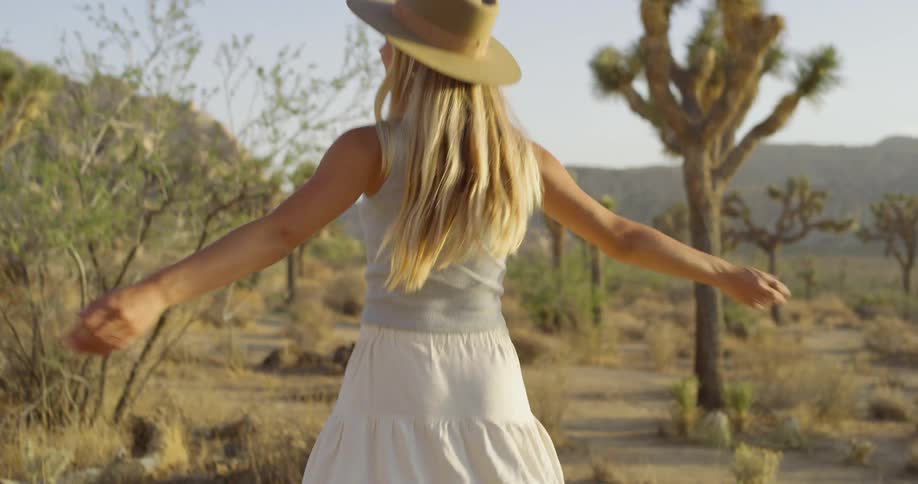 A blonde woman wearing a widebrimmed hat a light blue tank top a white skirt and brown cowboy boots walks through a desert landscape the background includes dry grass cacti and mountains under a bright sky