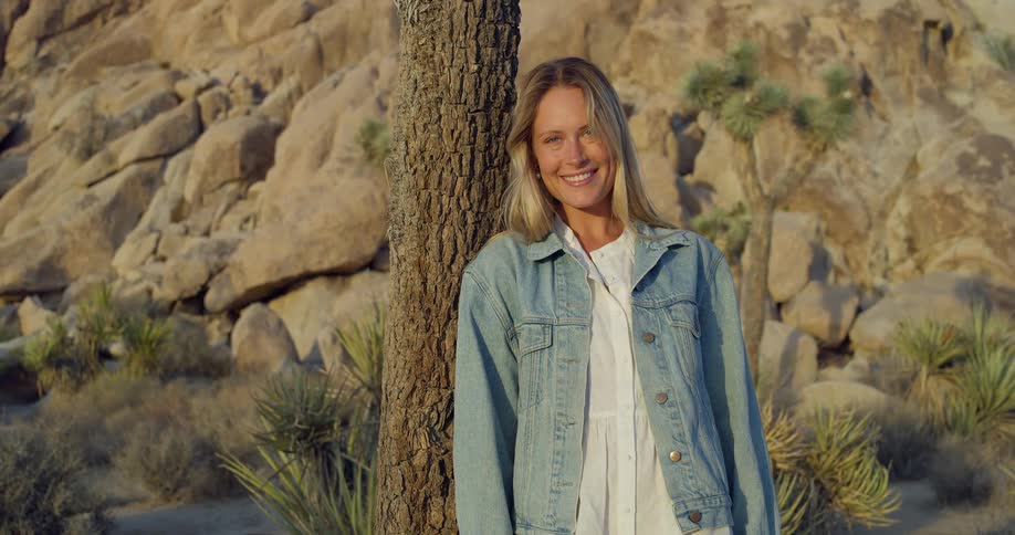 Blonde woman smiles leaning against a tree in the rocky desert