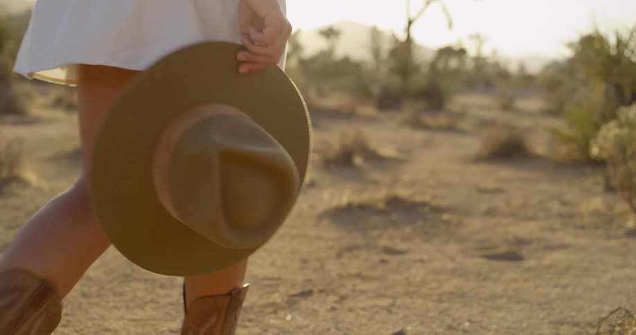 Female in a white skirt brown cowboys boots and carrying a western hat walking in the desert