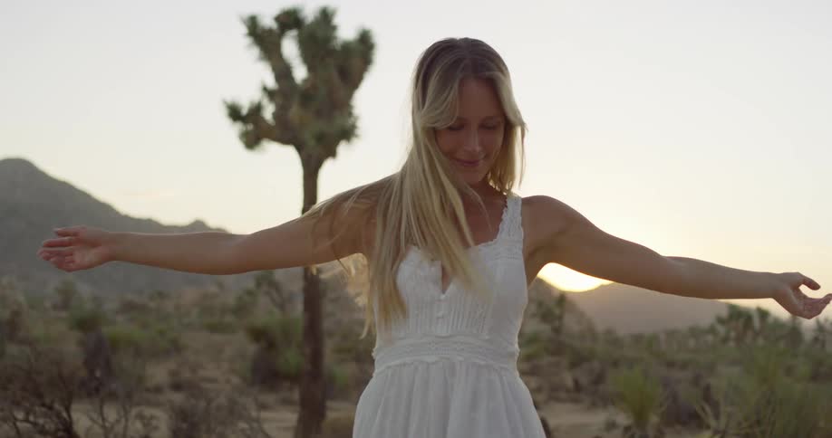 A young woman outdoors in the wild wearing a white gown and a gold necklace with long blonde hair looking happy as she spins around