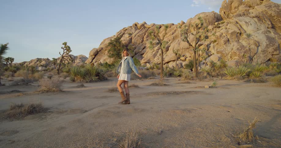 A wide shot shows a young woman walking through a desert landscape with large boulders and joshua trees she is wearing a white dress a blue denim jacket and brown cowboy boots