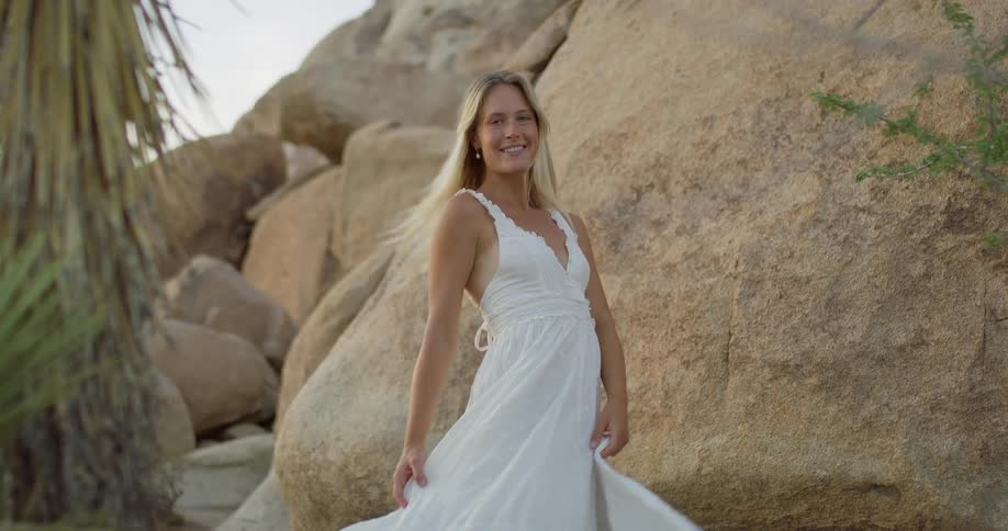 A woman with long blonde hair and a white dress stands in a desert with her back to the camera looking over her shoulder and smiling at a collection of large round rocks