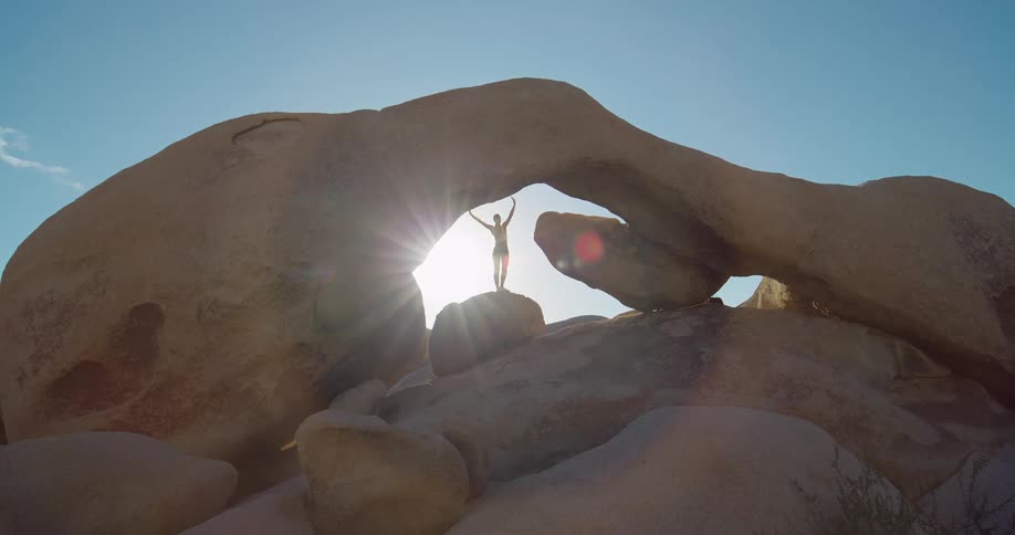 Person practices yoga in a balancing pose under a rock formation lit by the sun