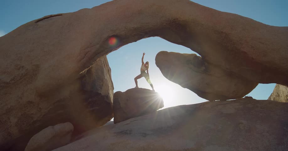 Person practices yoga on a rock beneath a natural arch lit by the sun