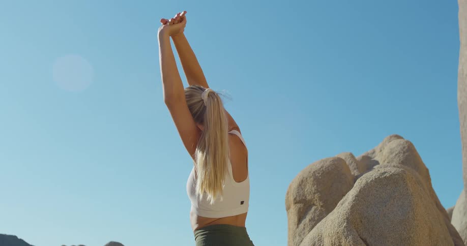 Woman in a sports top stretching her arms outdoors in a rocky area under a blue sky