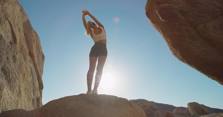Blonde woman stretches while standing on a rock formation and facing the bright sun