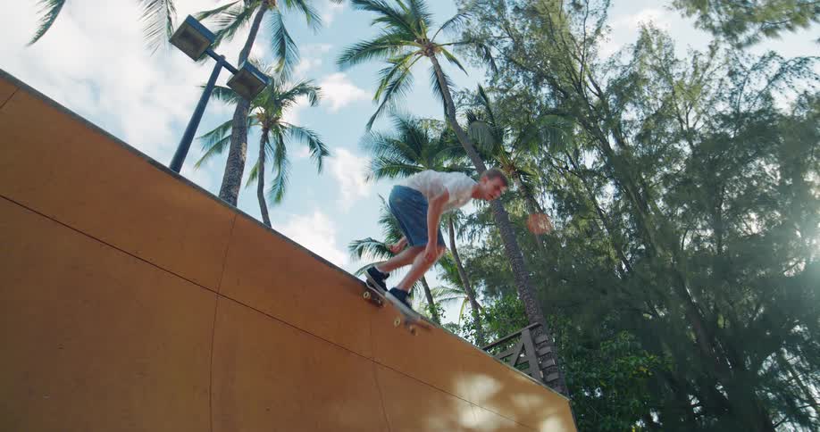 A skateboarder wearing a white shirt and blue shorts stands on the top edge of a wooden ramp balancing a skateboard on the coping before kicking the board down and dropping out of the frame