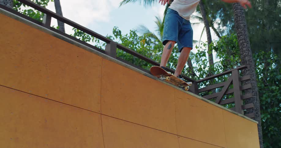 A lowangle shot shows a skateboarder on the top edge of a ramp the person is wearing jean shorts and black sneakers and is about to start their ride
