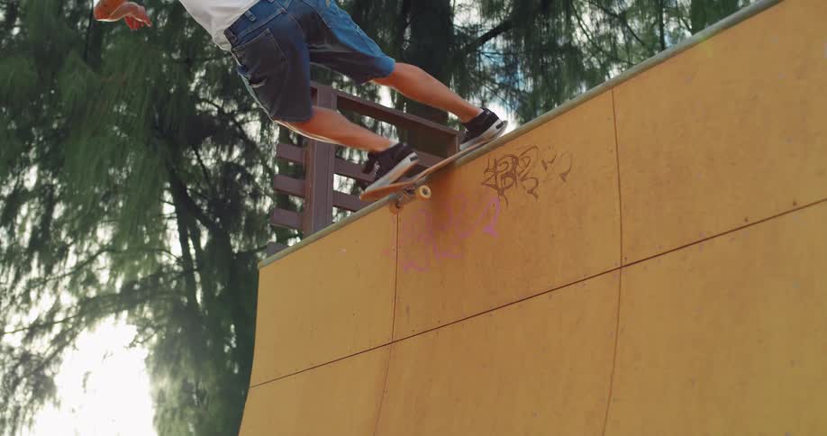 A skateboarder in a white shirt and blue shorts performs a trick on a wooden ramp with trees and a bright sky in the background