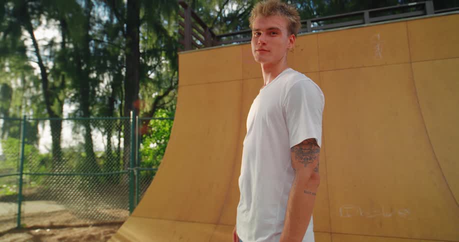 A skateboarder with a white shirt and jean shorts stands on the side of a wooden skate ramp with a skateboard on the ground near his feet facing the camera