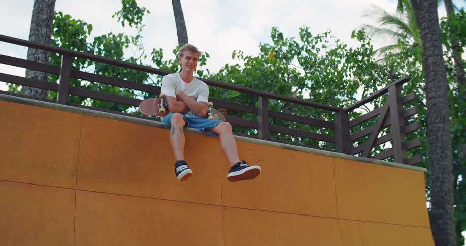 The man is sitting on the top of the ramp with his skateboard