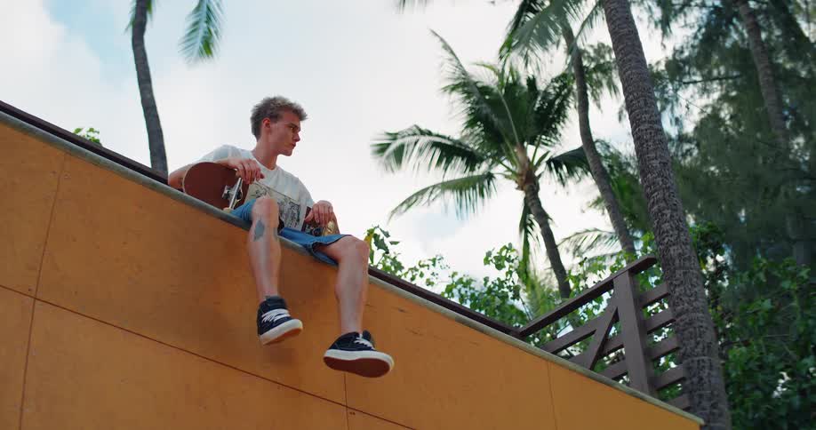 Young man sitting on top of a ramp holding a skateboard with palm trees in the background