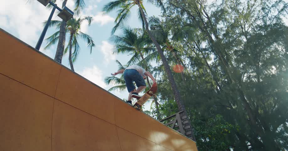 A skateboarder in a white shirt and dark blue shorts reaches the top of a wooden ramp balancing the skateboard on the coping before kicking the board down and riding off the right side of the frame