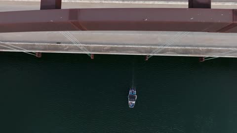 A series of cars and vans driving back and forth on a busy bridge with a boat passing underneath at midday