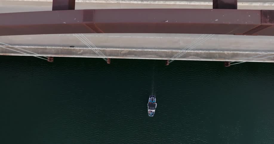 A series of cars and vans driving back and forth on a busy bridge with a boat passing underneath at midday