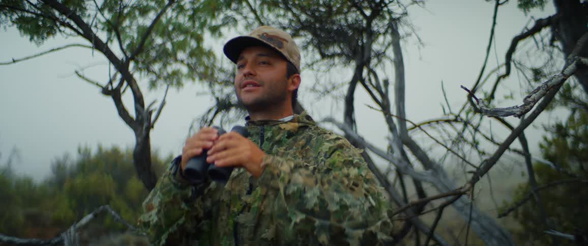 A man in camouflage gear and a cap raises a pair of binoculars to his eyes in a wooded area he then lowers the binoculars and looks up and to the side appearing to be scanning his surroundings