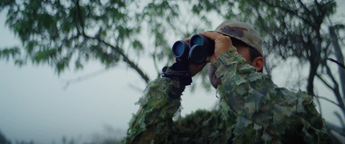 A person in camouflage gear and a cap looks through binoculars in a forest