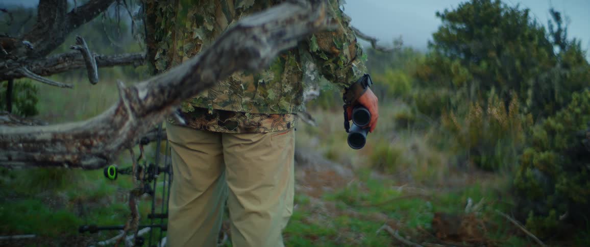 Person in camouflage clothing holds binoculars and a bow in a vegetated area