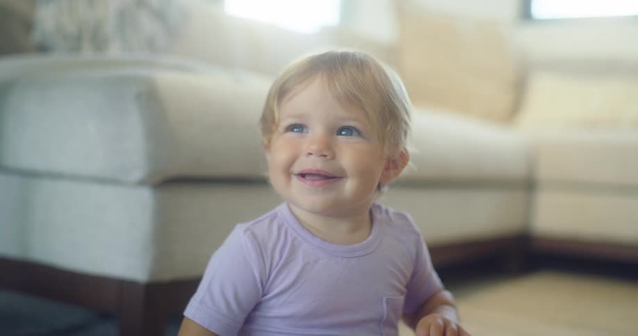 Blond baby in a lilac shirt smiles while looking up in a living room