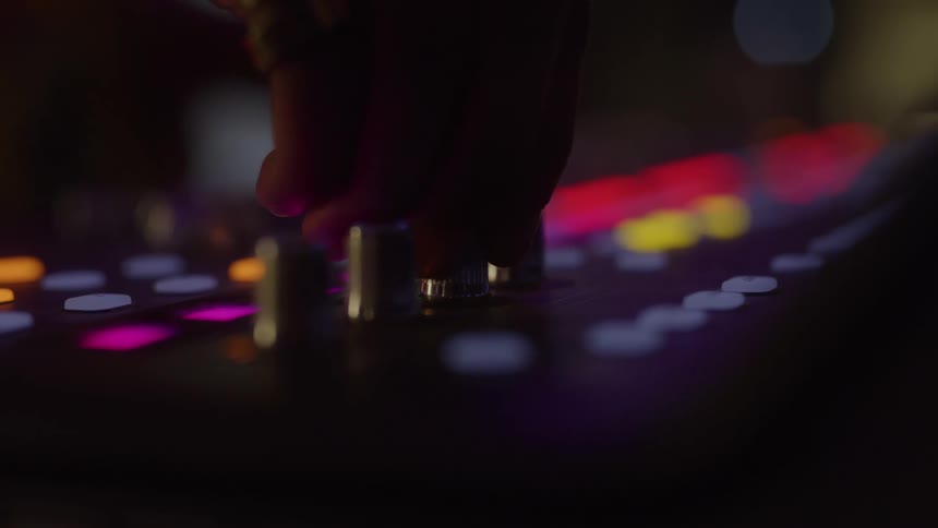 The video shows a closeup of a persons hand adjusting a dial on a sound mixing board the board is lit up with various colored lights