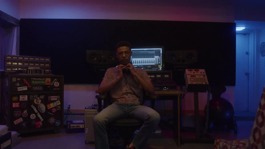 A person is seated in a dark music studio wearing glasses and a shortsleeved collared shirt they have headphones around their neck and are looking directly at the camera with a neutral expression