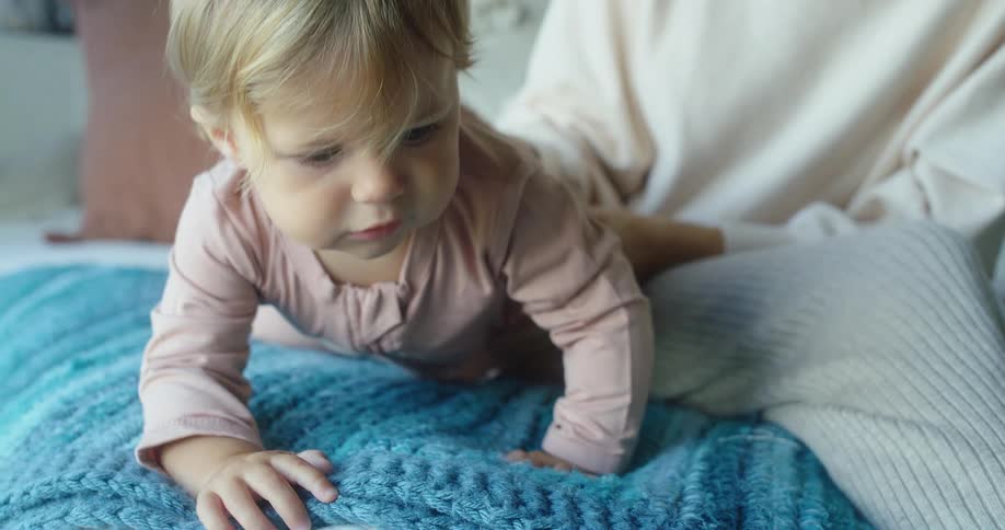 The video shows a woman with blonde hair wearing a light sweater sitting on a bed and smiling down at a baby the baby also with blonde hair is wearing a pink onesie and crawling on a blue blanket