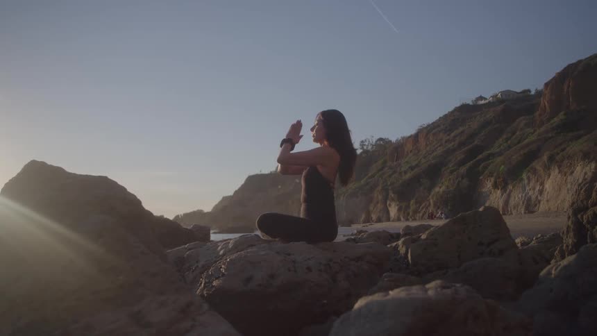 The video shows a woman performing a yoga pose on a beach she is sitting on a large rock with her legs crossed and she raises her arms into the air bringing her palms together in a prayer position the sun shines brightly behind her creating a silhoue