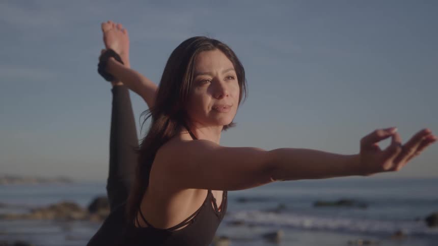 A woman with dark hair wearing a black top and dark pants is performing a yoga pose near the ocean with a rocky shoreline in the background her right hand holding her right foot behind her while her left arm is extended forward