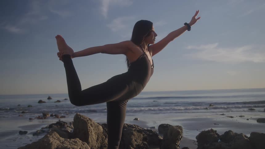 A woman in athletic wear performs a yoga pose on a rocky beach with the ocean and sky in the background