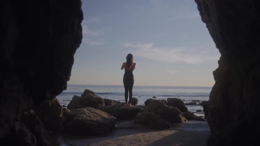 The video shows a wide shot of a woman performing a yoga pose on a large rock on a beach she is seen from a distance with her back to the camera framed between two large dark rock formations facing the calm ocean and the horizon