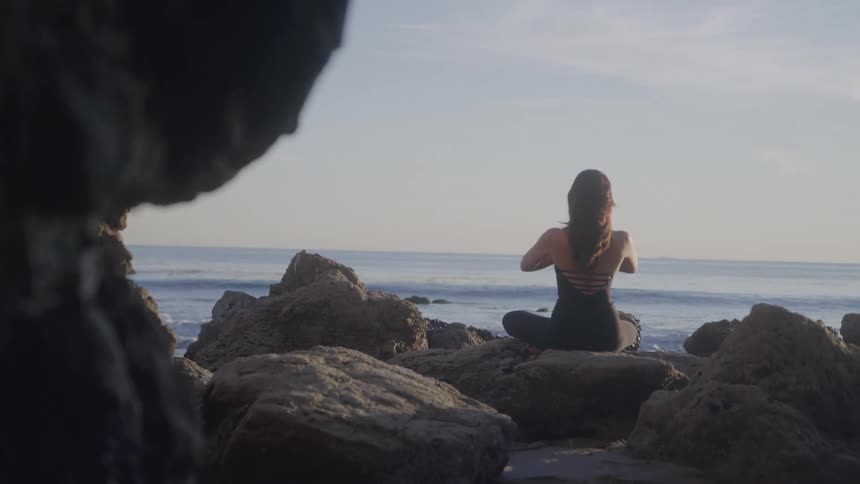 A woman practices yoga by the ocean on a rocky beach at sunrise