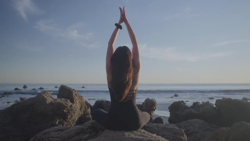 Woman does yoga on coastal rocks facing the quiet ocean
