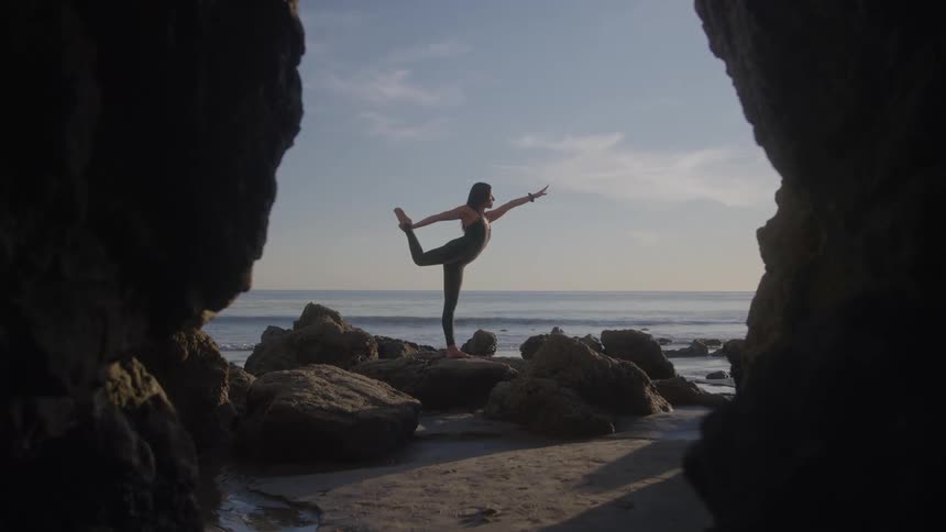 A woman practices yoga on a rocky beach by the ocean cave