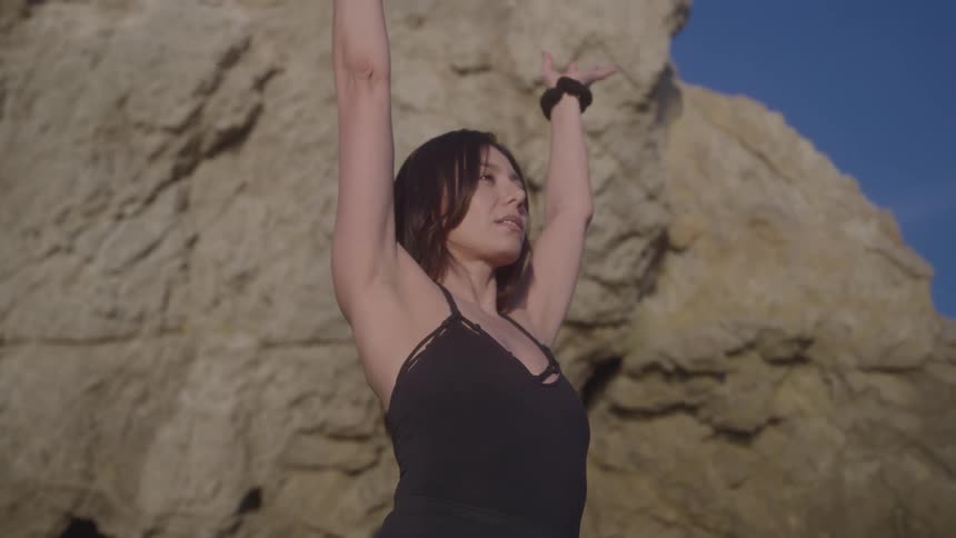 A woman is seen from the chest up with her arms raised above her head performing a stretching exercise against the backdrop of a large rocky cliff on a bright day