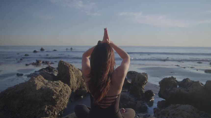 Woman stretches and sits in a yoga pose amongst the boulders on the coast on a cloudy day