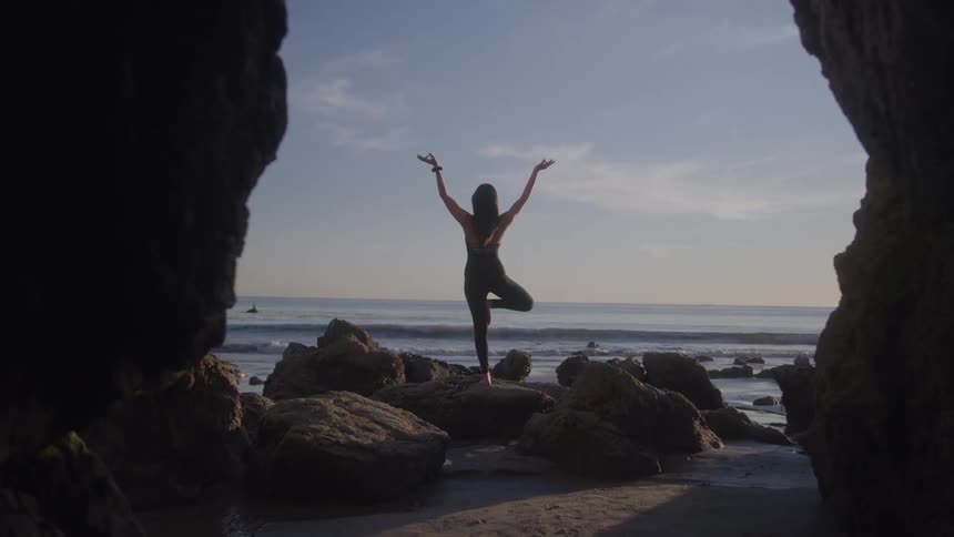 Person practicing a yoga pose standing on a rock by the sea viewed from inside a cave