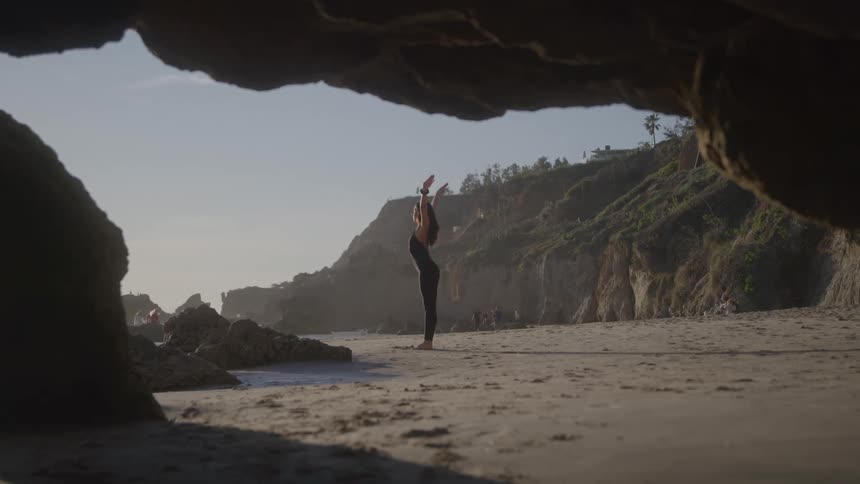 A woman practices yoga on a beach under a rocky cave with ocean views