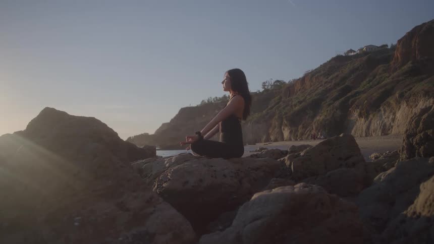 Woman meditates on coastal rocks at sunset