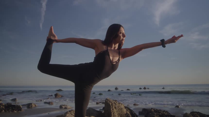 A woman performing a yoga pose on rocks by the ocean during sunset