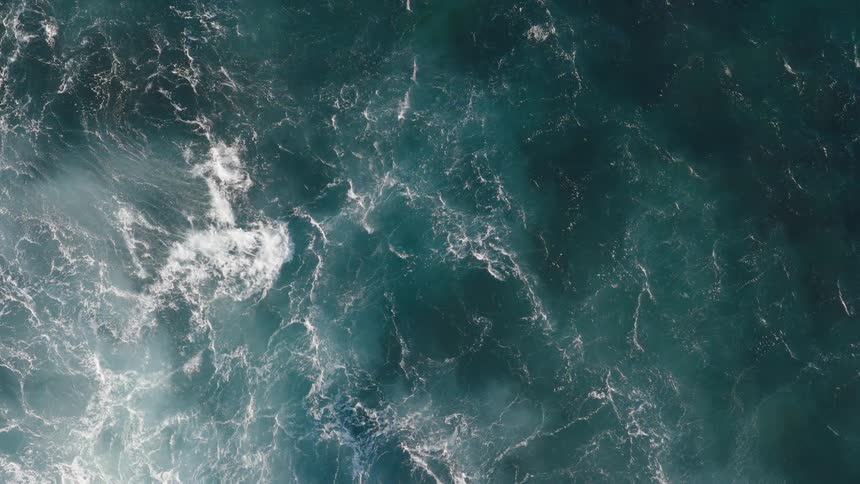 A top down view shows the rough surface of a dark blue ocean with bright white foam from the waves slowly rolling and swirling over the water