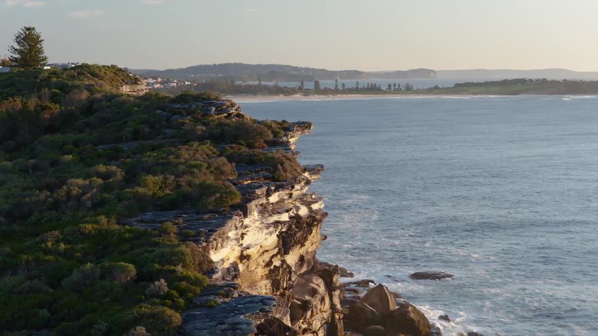 A rugged bush covered cliff overlooks the ocean with a distant town and coastline visible in the soft light of sunrise or sunset